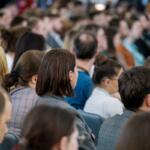 Crowd of attendees seated closely at an indoor conference, viewed from behind.