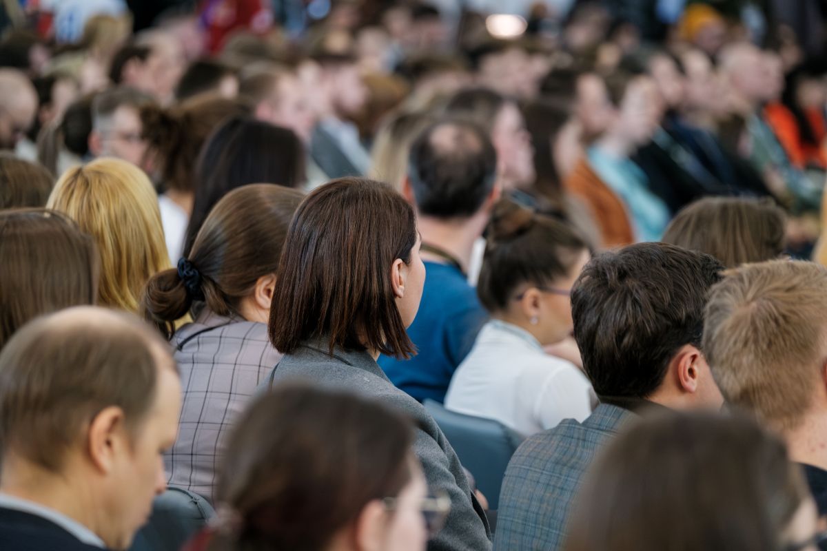 Crowd of attendees seated closely at an indoor conference, viewed from behind.