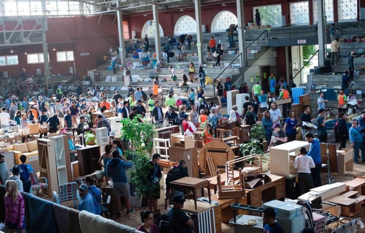 Busy indoor flea market with shoppers browsing secondhand furniture and tables scattered across the floor.