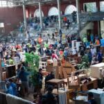 Busy indoor flea market with shoppers browsing secondhand furniture and tables scattered across the floor.