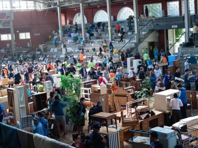 Busy indoor flea market with shoppers browsing secondhand furniture and tables scattered across the floor.