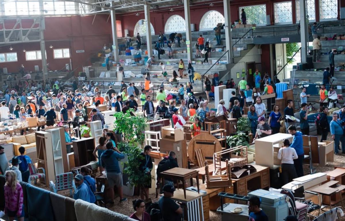 Busy indoor flea market with shoppers browsing secondhand furniture and tables scattered across the floor.