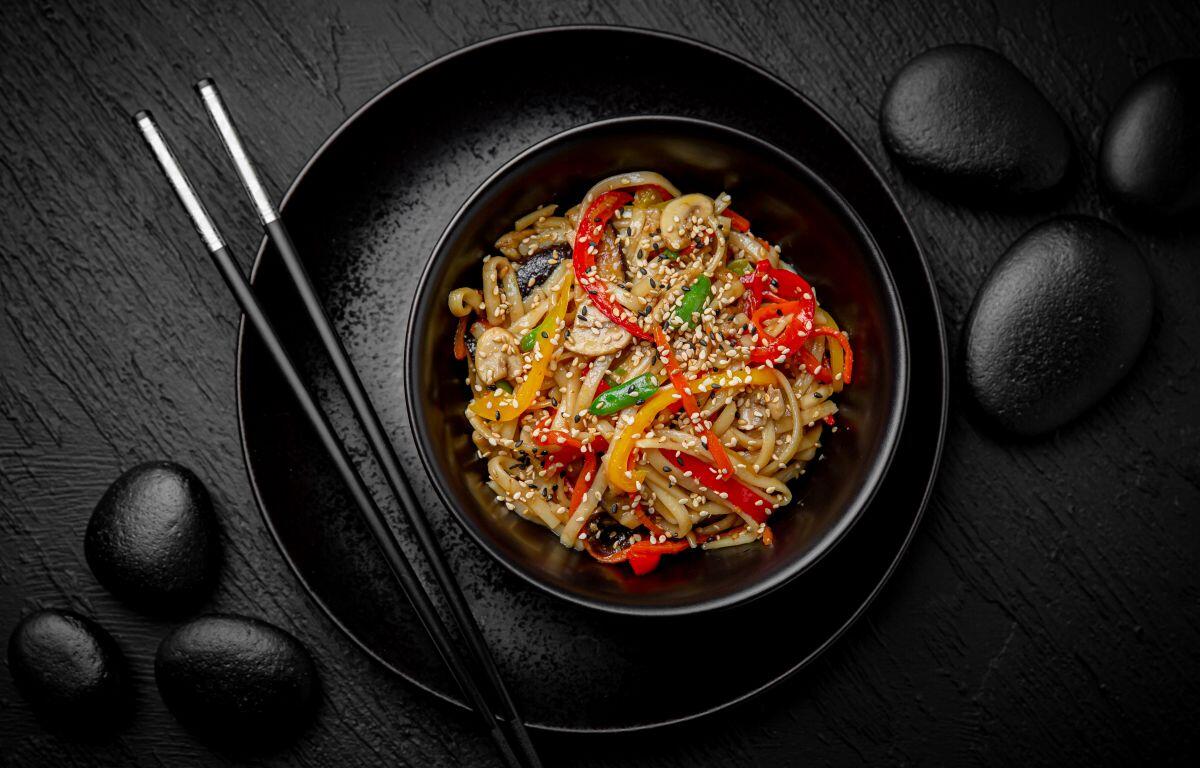 Bowl of vegetable noodles with sesame seeds on a black plate, chopsticks resting beside, dark stone background.