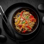 Bowl of vegetable noodles with sesame seeds on a black plate, chopsticks resting beside, dark stone background.