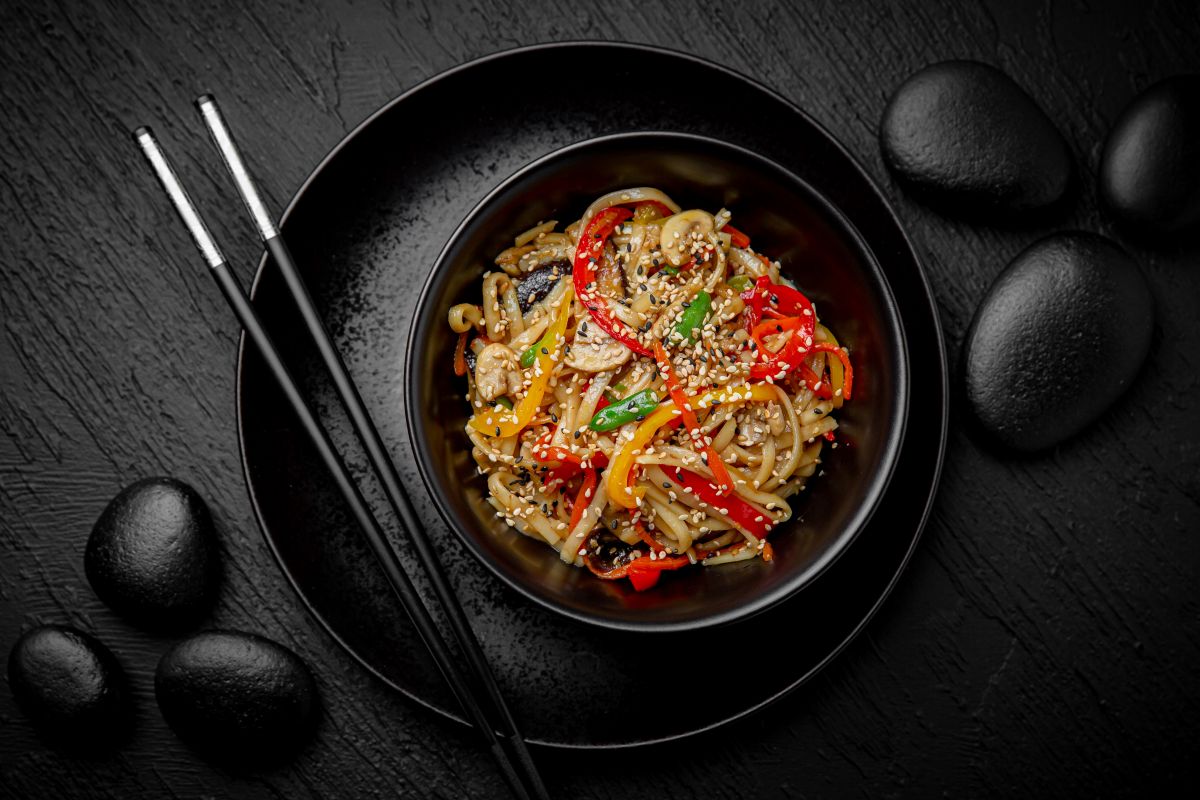 Bowl of vegetable noodles with sesame seeds on a black plate, chopsticks resting beside, dark stone background.