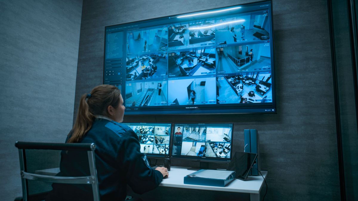 Security operator monitoring multiple CCTV feeds on a wall-sized monitor in a control room.