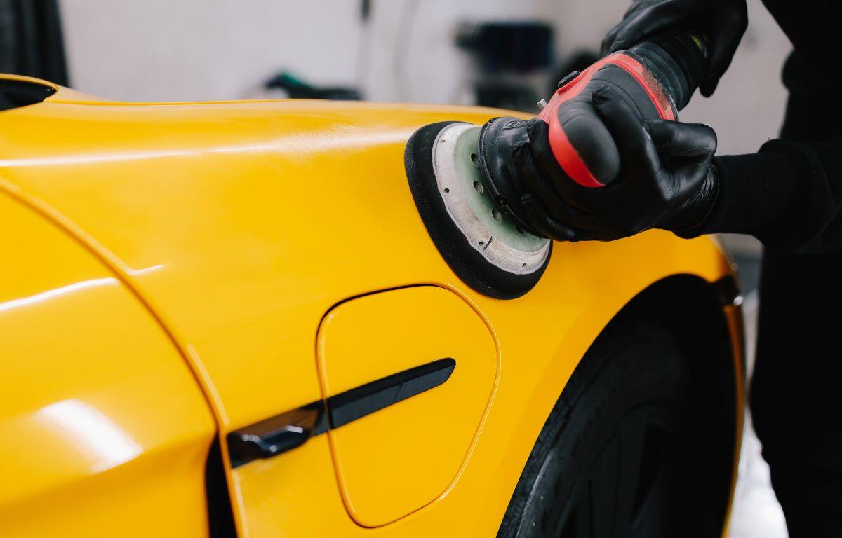 Person wearing gloves polishes a bright yellow car with a polishing machine (buffer).