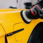 Person wearing gloves polishes a bright yellow car with a polishing machine (buffer).