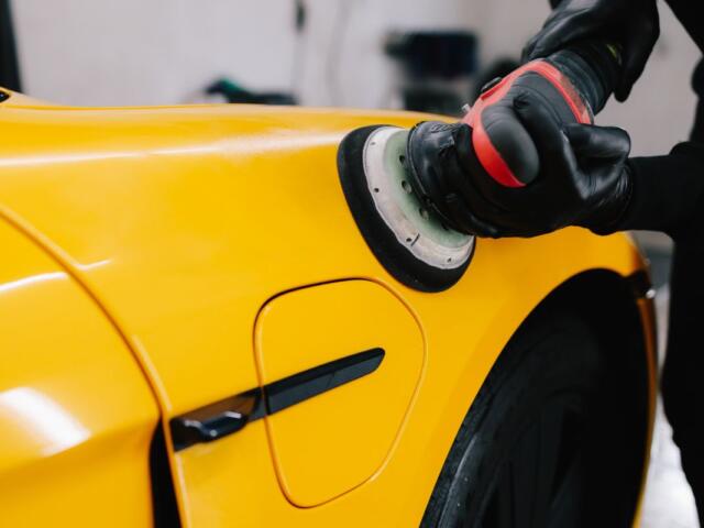 Person wearing gloves polishes a bright yellow car with a polishing machine (buffer).