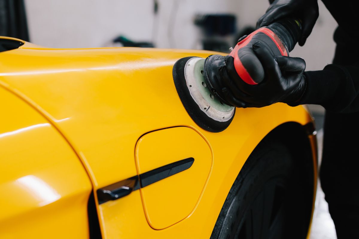 Person wearing gloves polishes a bright yellow car with a polishing machine (buffer).