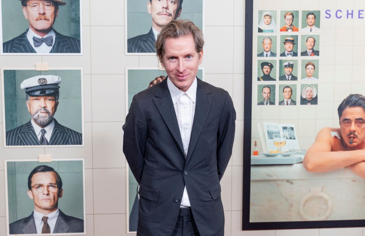 Man in a dark suit stands with hands behind his back in front of a wall displaying framed portrait photos of men in formal attire.
