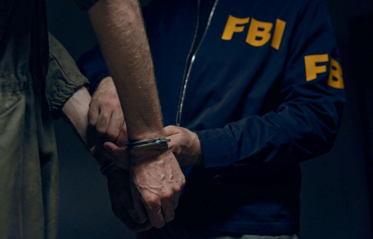 Close-up of hands in handcuffs being secured by an FBI agent in a blue jacket with yellow 'FBI' lettering behind him.