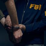 Close-up of hands in handcuffs being secured by an FBI agent in a blue jacket with yellow 'FBI' lettering behind him.