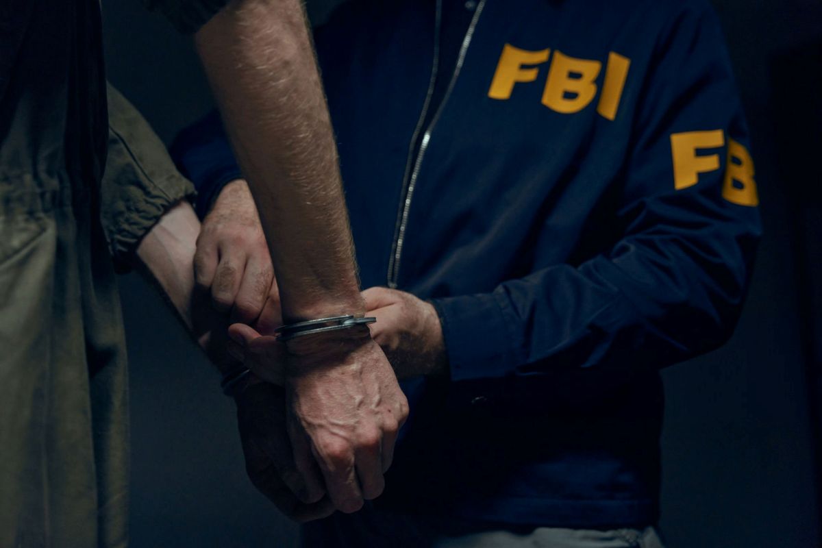 Close-up of hands in handcuffs being secured by an FBI agent in a blue jacket with yellow 'FBI' lettering behind him.