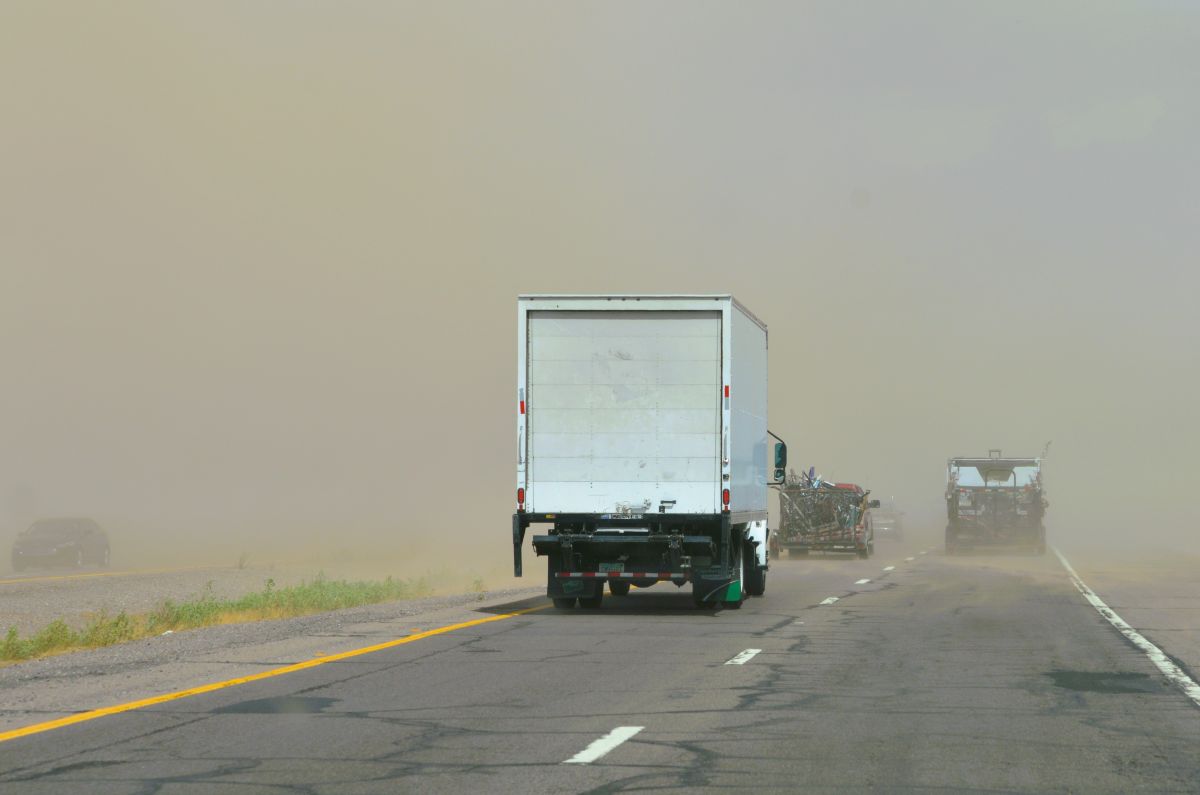 White box truck on a dusty highway with other vehicles following in a sandstorm, limited visibility.