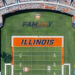 Aerial view of an Illinois football stadium with the field showing a large orange 'ILLINOIS' end zone and surrounding orange seating accents.