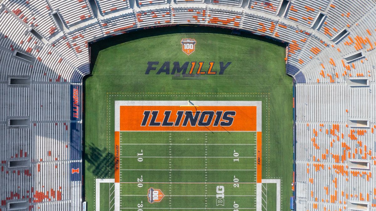 Aerial view of an Illinois football stadium with the field showing a large orange 'ILLINOIS' end zone and surrounding orange seating accents.