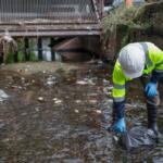 Worker in a neon safety jacket and hard hat collects a plastic bag from a polluted drainage pool under a metal bridge.