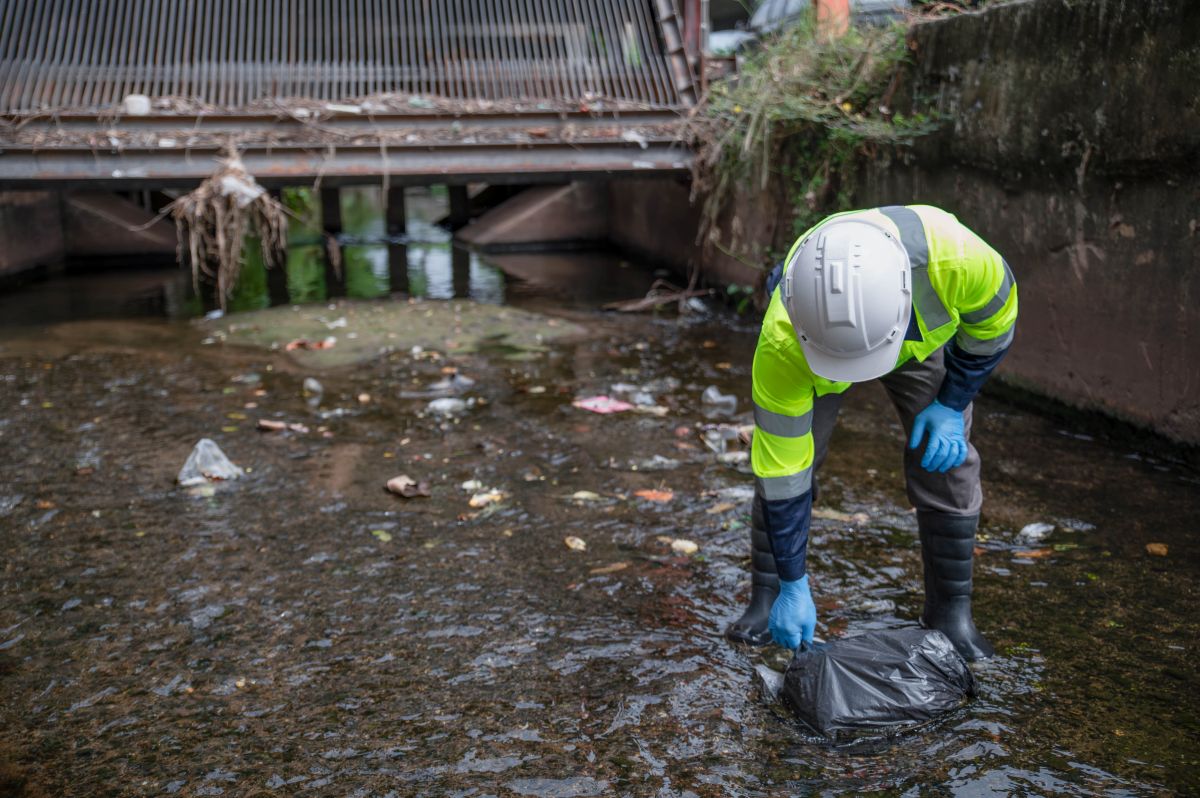 Worker in a neon safety jacket and hard hat collects a plastic bag from a polluted drainage pool under a metal bridge.