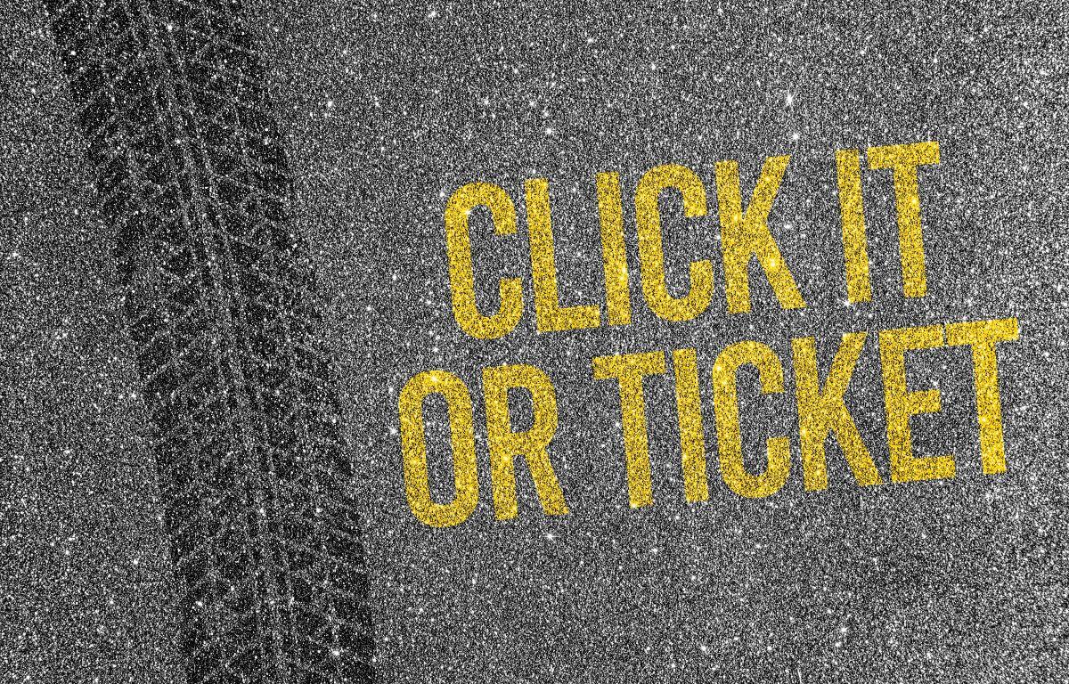 Yellow stenciled message 'CLICK IT OR TICKET' on an asphalt road beside a tire track.