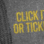 Yellow stenciled message 'CLICK IT OR TICKET' on an asphalt road beside a tire track.