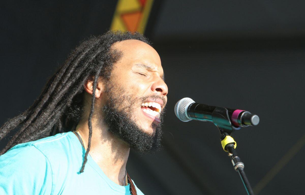 Male singer with long dreadlocks and beard singing with eyes closed into a microphone on stage in a turquoise shirt on a sunny performance.