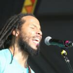 Male singer with long dreadlocks and beard singing with eyes closed into a microphone on stage in a turquoise shirt on a sunny performance.