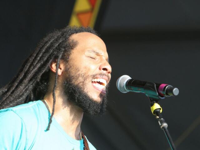 Male singer with long dreadlocks and beard singing with eyes closed into a microphone on stage in a turquoise shirt on a sunny performance.