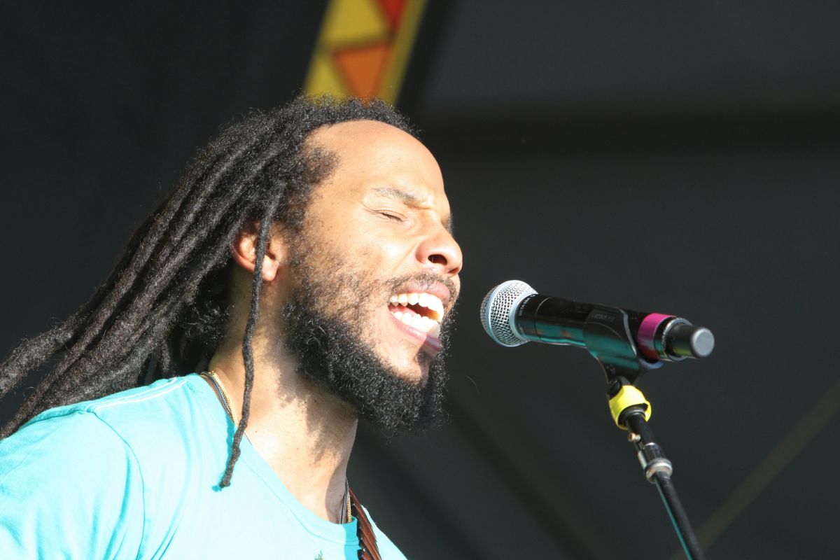 Male singer with long dreadlocks and beard singing with eyes closed into a microphone on stage in a turquoise shirt on a sunny performance.