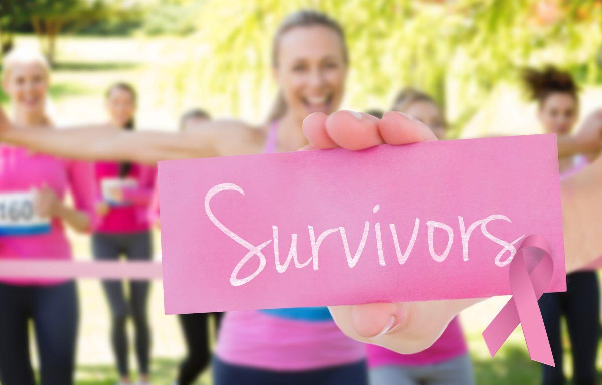 Group of women in pink exercise outfits holding a pink card that says 'Survivors' at a park, pink ribbon visible for breast cancer awareness