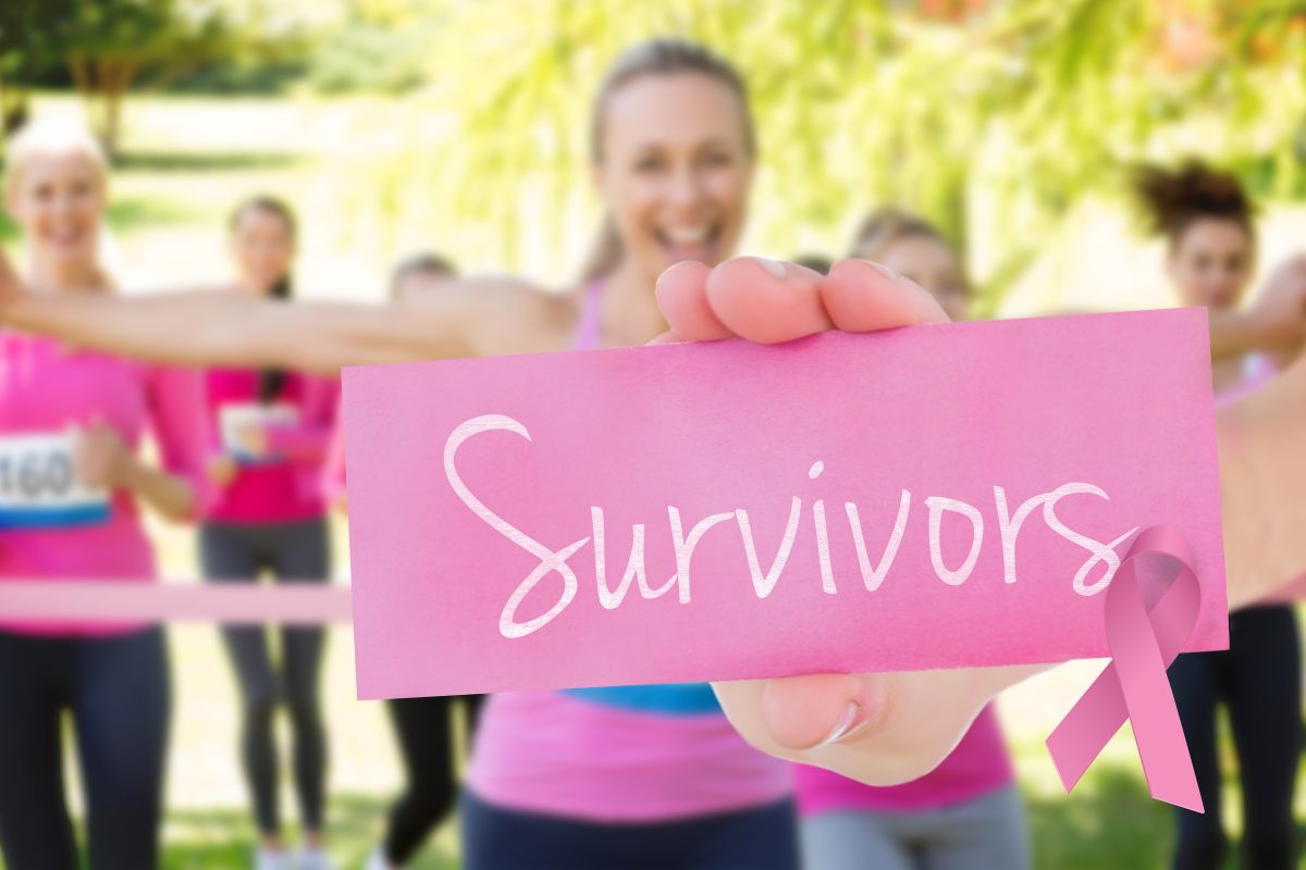 Group of women in pink exercise outfits holding a pink card that says 'Survivors' at a park, pink ribbon visible for breast cancer awareness