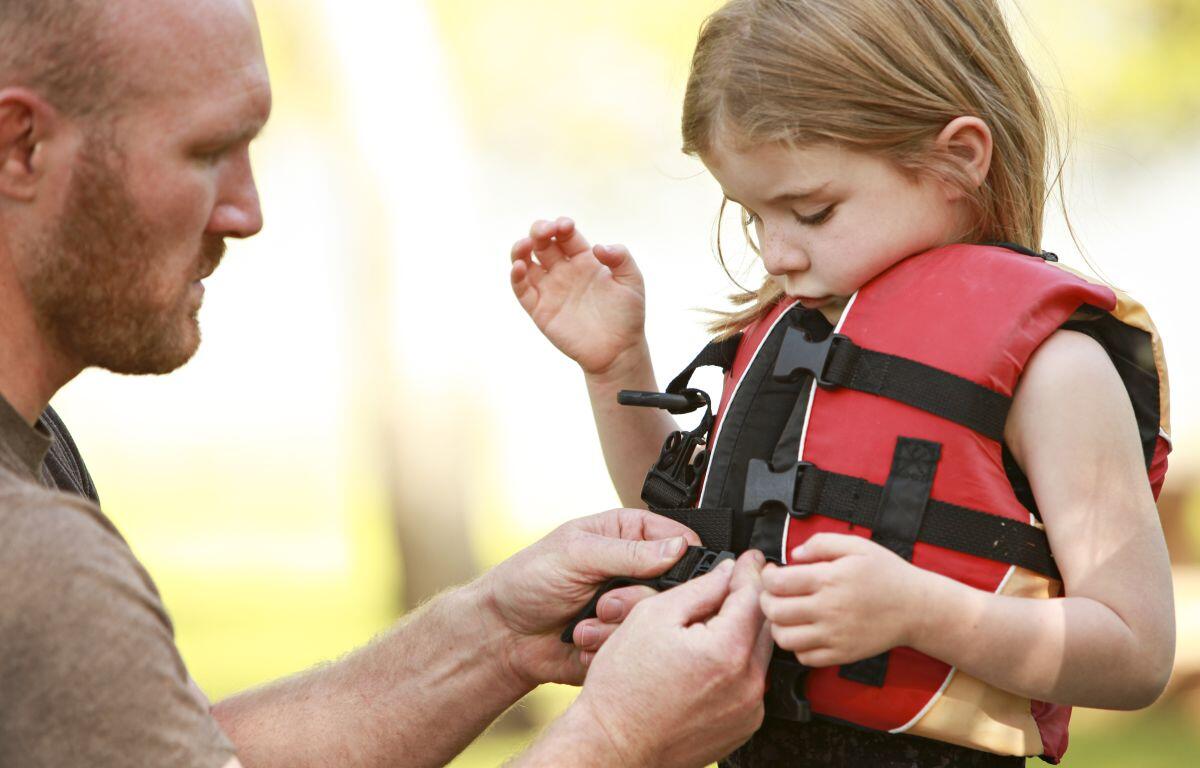 Man helping a young girl fasten a red life jacket outdoors.
