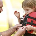 Man helping a young girl fasten a red life jacket outdoors.