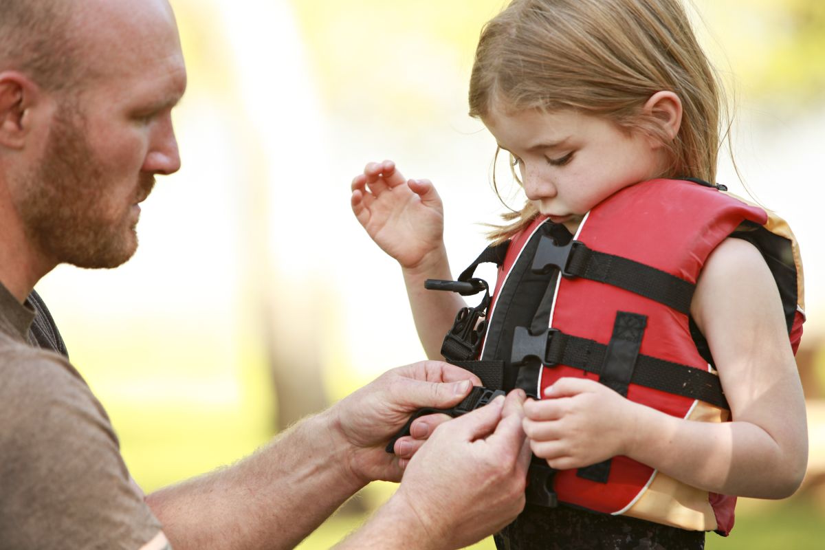 Man helping a young girl fasten a red life jacket outdoors.
