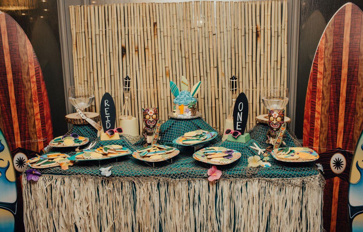 Tiki-themed dessert table with bamboo backdrop, surfboard decorations at sides, netting, and tropical treats including a turquoise cake and cookie plates.