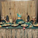 Tiki-themed dessert table with bamboo backdrop, surfboard decorations at sides, netting, and tropical treats including a turquoise cake and cookie plates.