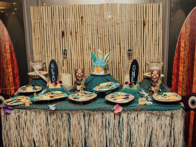 Tiki-themed dessert table with bamboo backdrop, surfboard decorations at sides, netting, and tropical treats including a turquoise cake and cookie plates.