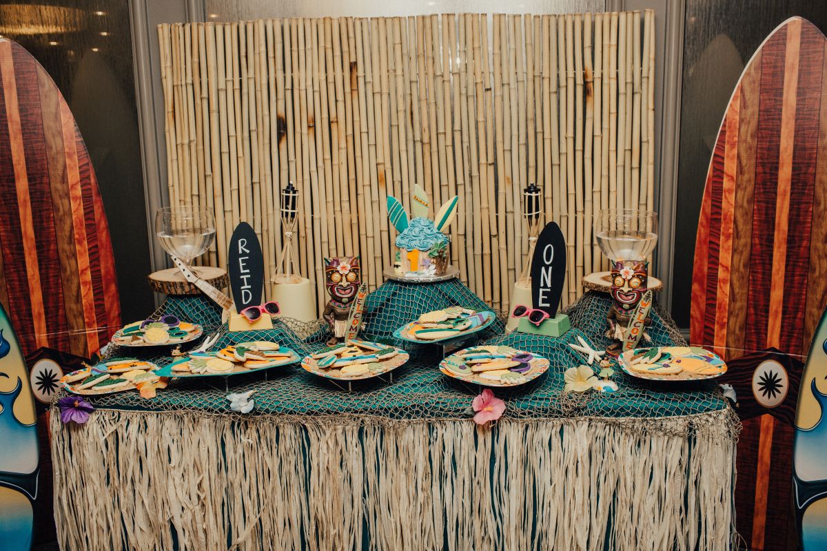 Tiki-themed dessert table with bamboo backdrop, surfboard decorations at sides, netting, and tropical treats including a turquoise cake and cookie plates.