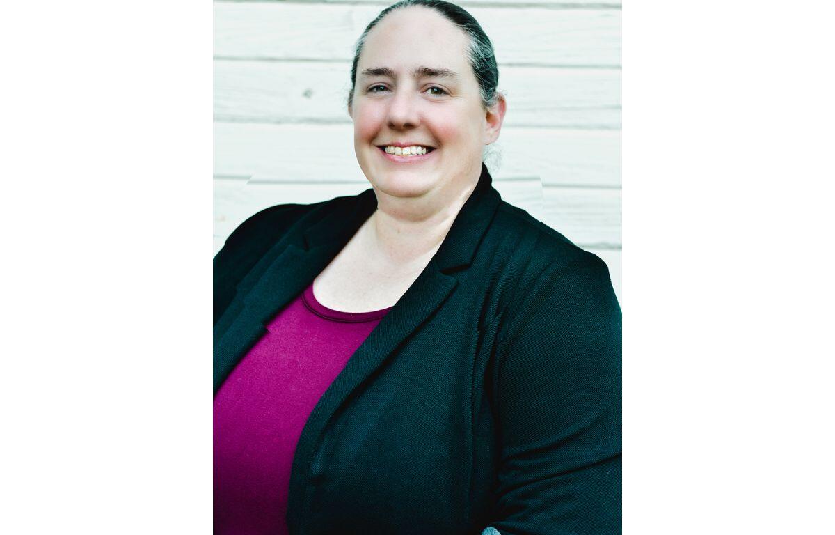 Smiling woman in a black blazer and magenta top, posed against a pale wooden backdrop.