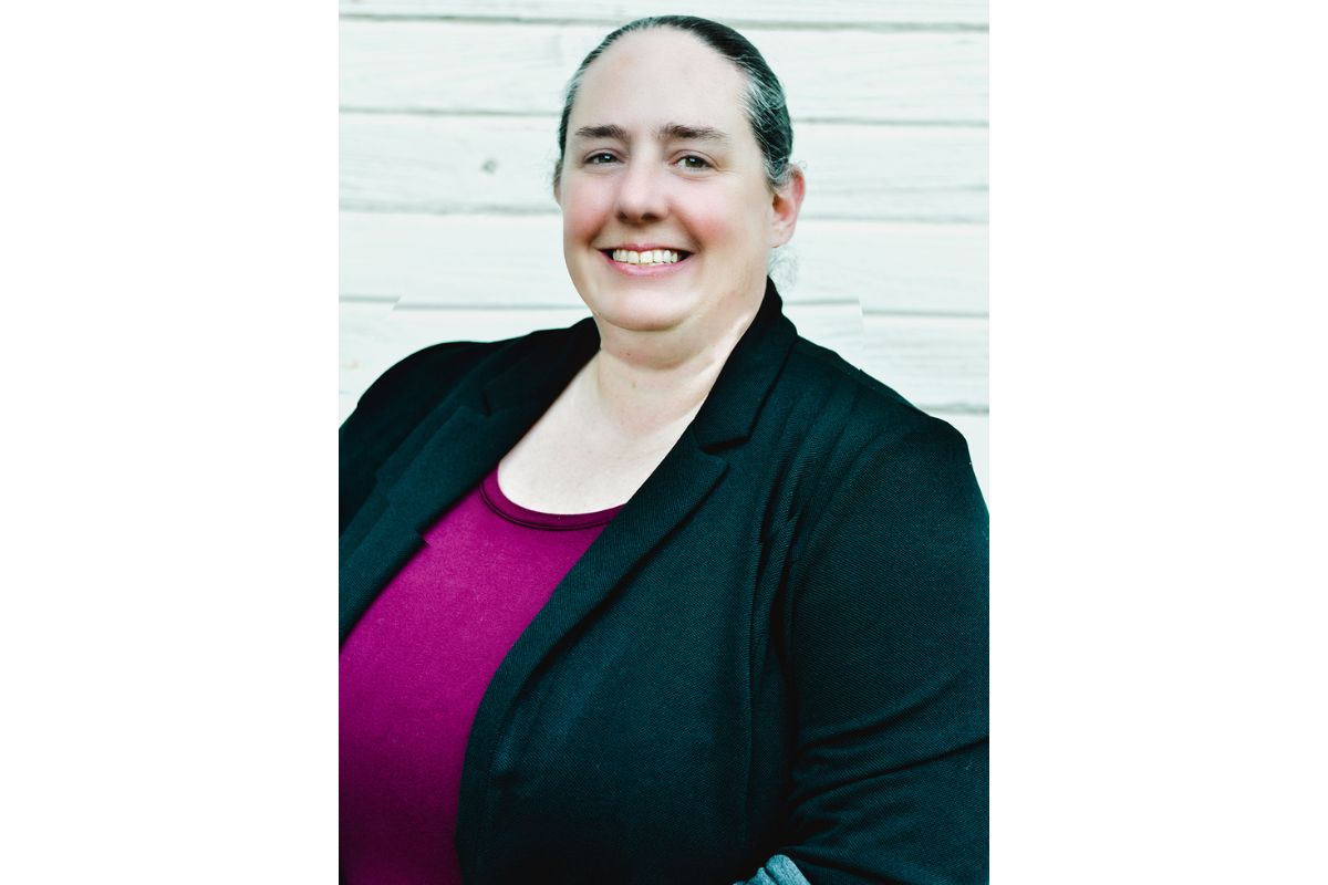 Smiling woman in a black blazer and magenta top, posed against a pale wooden backdrop.