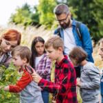 Group of children with a bearded man looking at plants outdoors as one child uses a magnifying glass to inspect leaves.