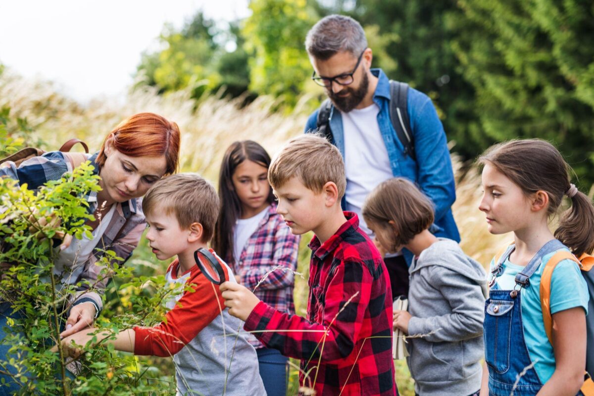 Group of children with a bearded man looking at plants outdoors as one child uses a magnifying glass to inspect leaves.