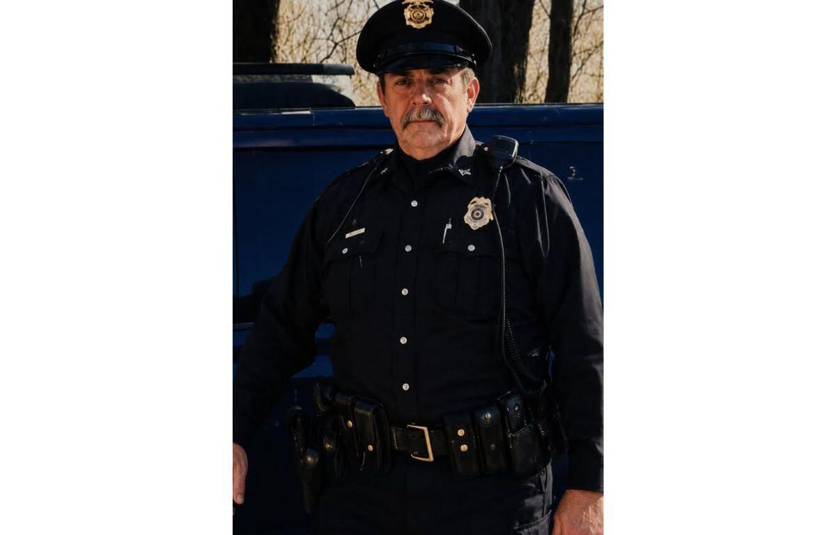 Police officer in uniform standing beside a dark vehicle outdoors, badge visible on chest and hat
