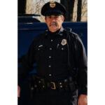 Police officer in uniform standing beside a dark vehicle outdoors, badge visible on chest and hat