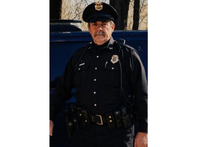 Police officer in uniform standing beside a dark vehicle outdoors, badge visible on chest and hat