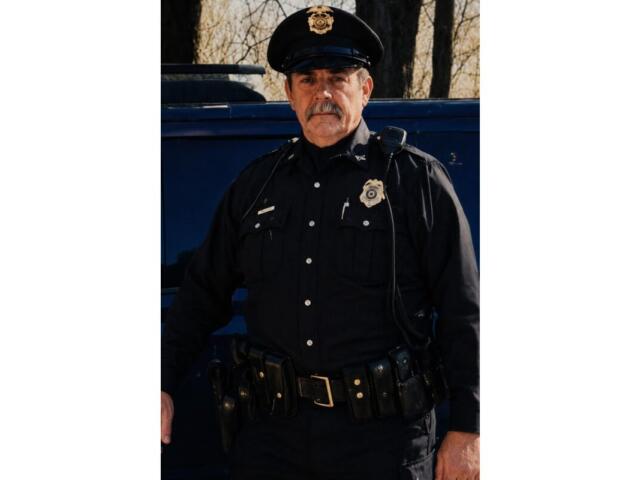 Police officer in a dark uniform with badge, standing beside a blue squad car outdoors.