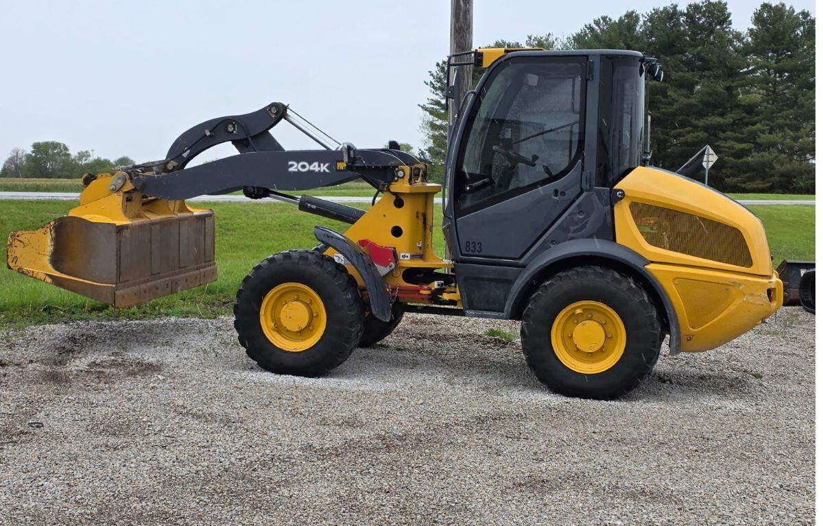Side view of a yellow compact wheel loader with a large front bucket raised slightly, parked on a gravel surface by a grassy field and road in the background.