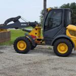 Side view of a yellow compact wheel loader with a large front bucket raised slightly, parked on a gravel surface by a grassy field and road in the background.