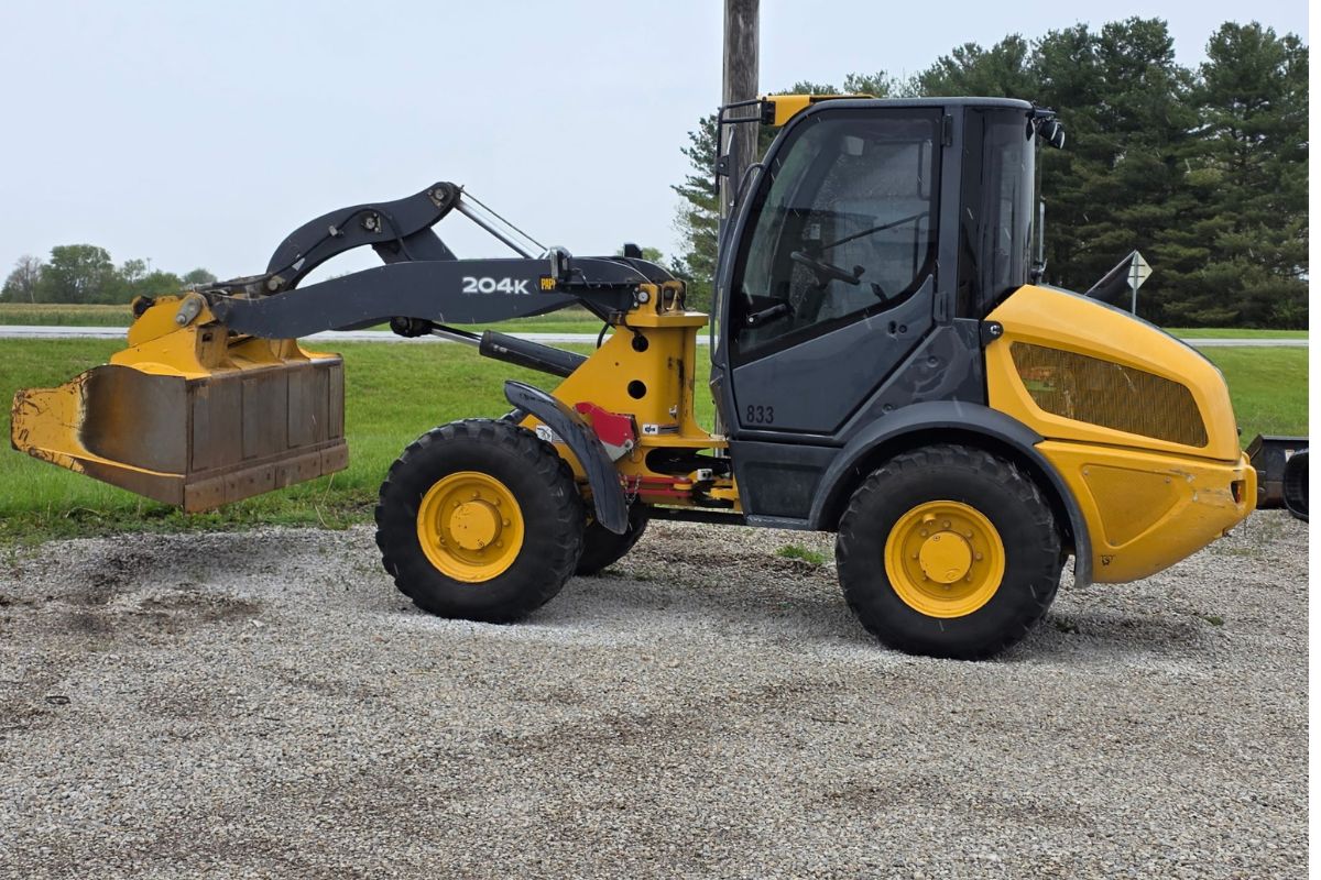 Side view of a yellow compact wheel loader with a large front bucket raised slightly, parked on a gravel surface by a grassy field and road in the background.
