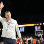 Illinois Fighting Illini coach waves to the crowd on a basketball court.